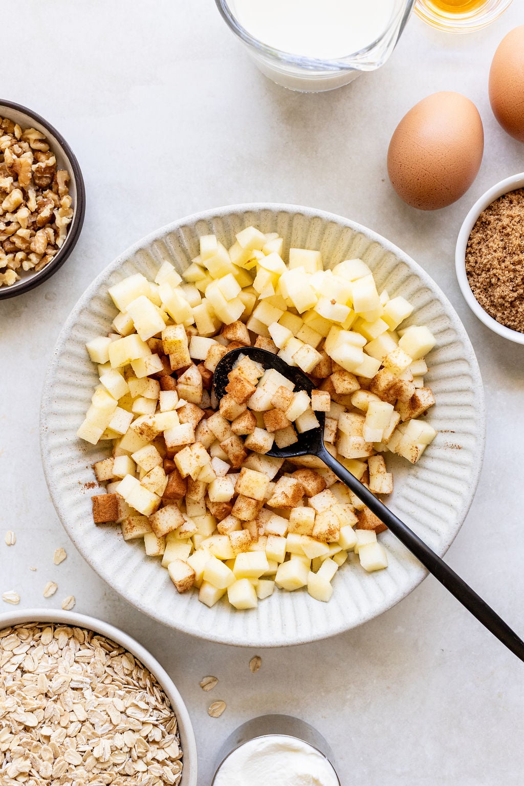 Diced apples with cinnamon in a bowl with a spoon, surrounded by oats, walnuts, eggs, brown sugar, milk, and yogurt on a light surface.
