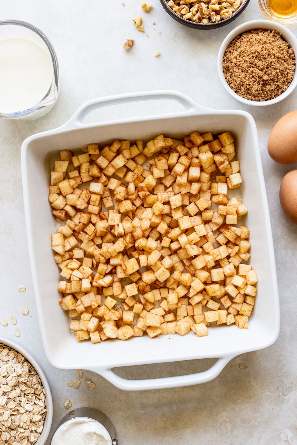 A baking dish filled with diced apples coated in cinnamon, surrounded by ingredients including eggs, milk, brown sugar, oats, walnuts, and flour on a light surface.