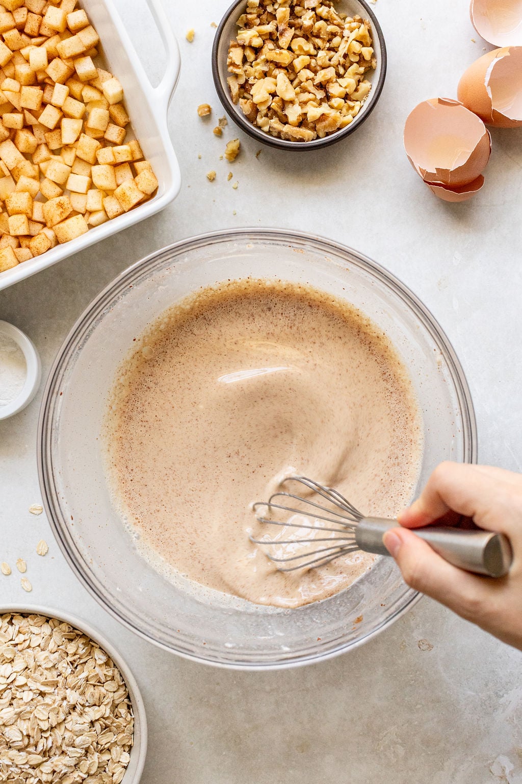 A hand whisks a creamy mixture in a glass bowl surrounded by chopped apples, walnuts, eggshells, oats, and a baking dish on a light countertop.