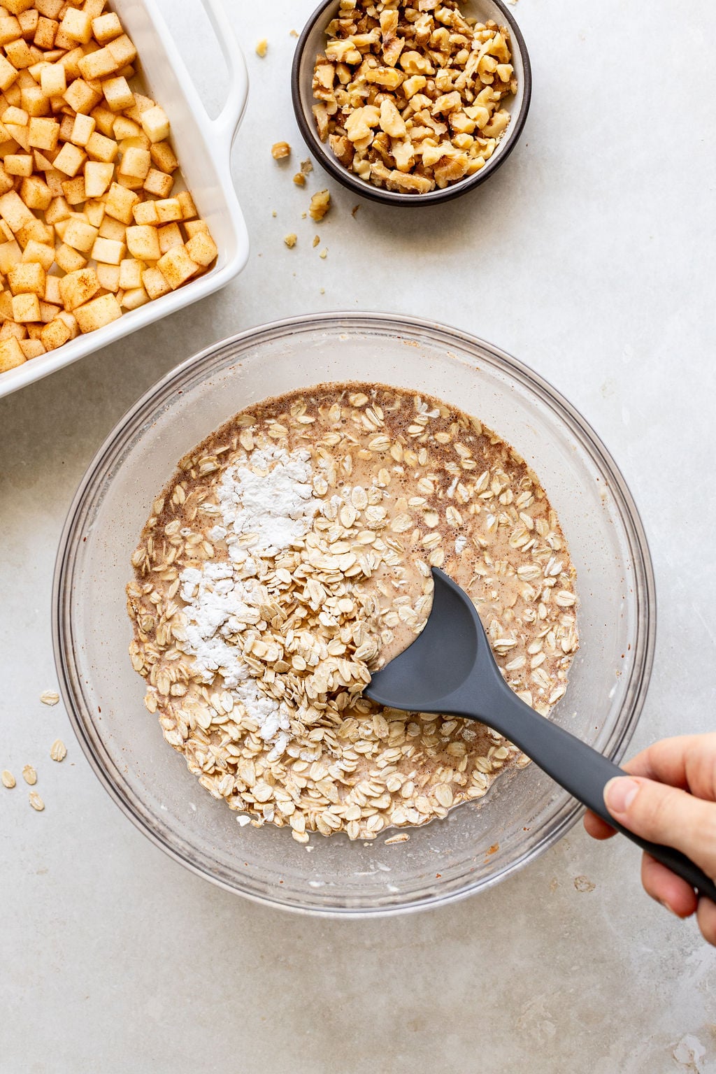 A hand stirs oats, flour, and liquid in a glass bowl with a black spoon.