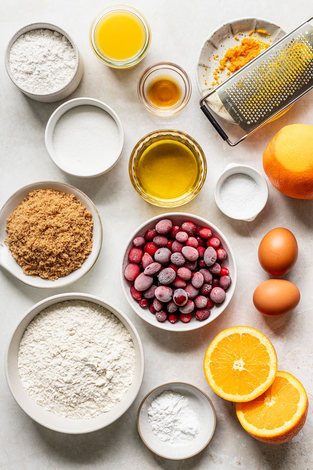 Assorted baking ingredients including flour, sugar, brown sugar, eggs, butter, oil, orange, orange zest, frozen cranberries, baking powder, and vanilla, arranged on a white surface.