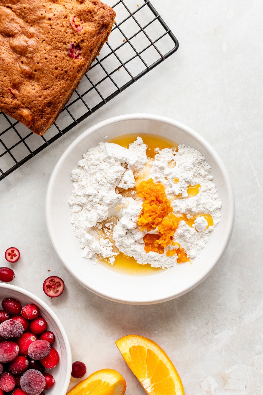 Bowl with powdered sugar, orange zest, and juice next to a wire rack with baked bread, sliced oranges, and cranberries on a light surface.