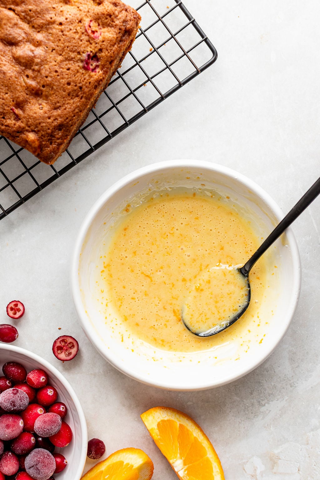 A bowl of yellow glaze with a spoon, next to a bowl of cranberries, orange slices, and a loaf of bread on a cooling rack.