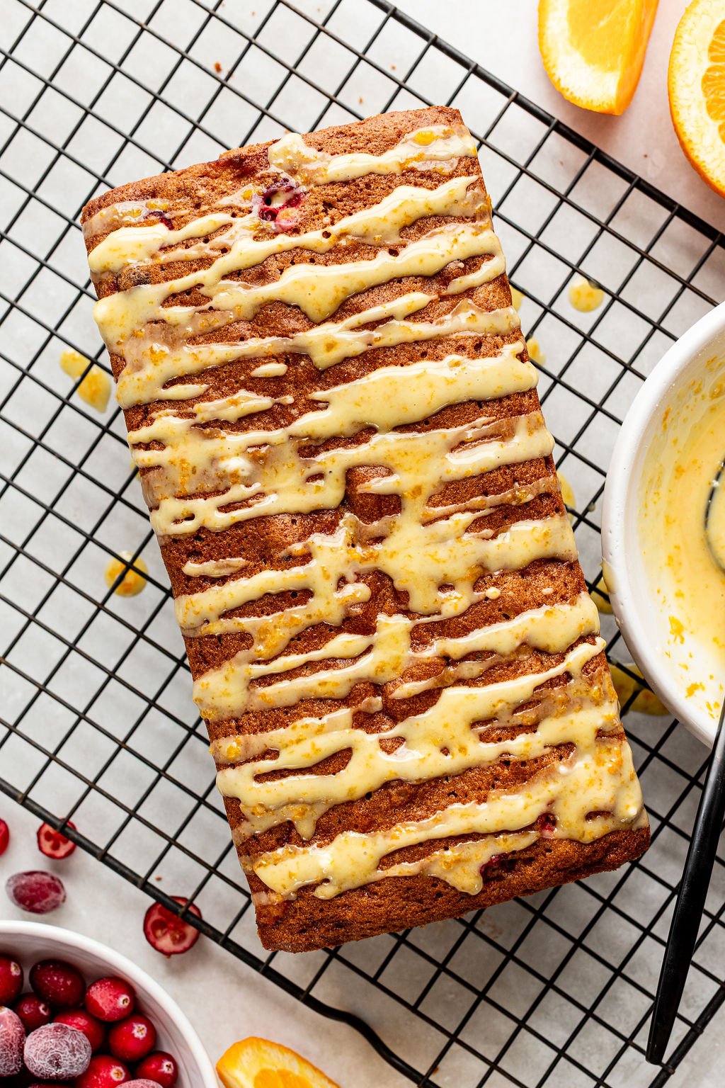 A loaf of bread topped with orange glaze sits on a cooling rack, surrounded by orange slices and cranberries.