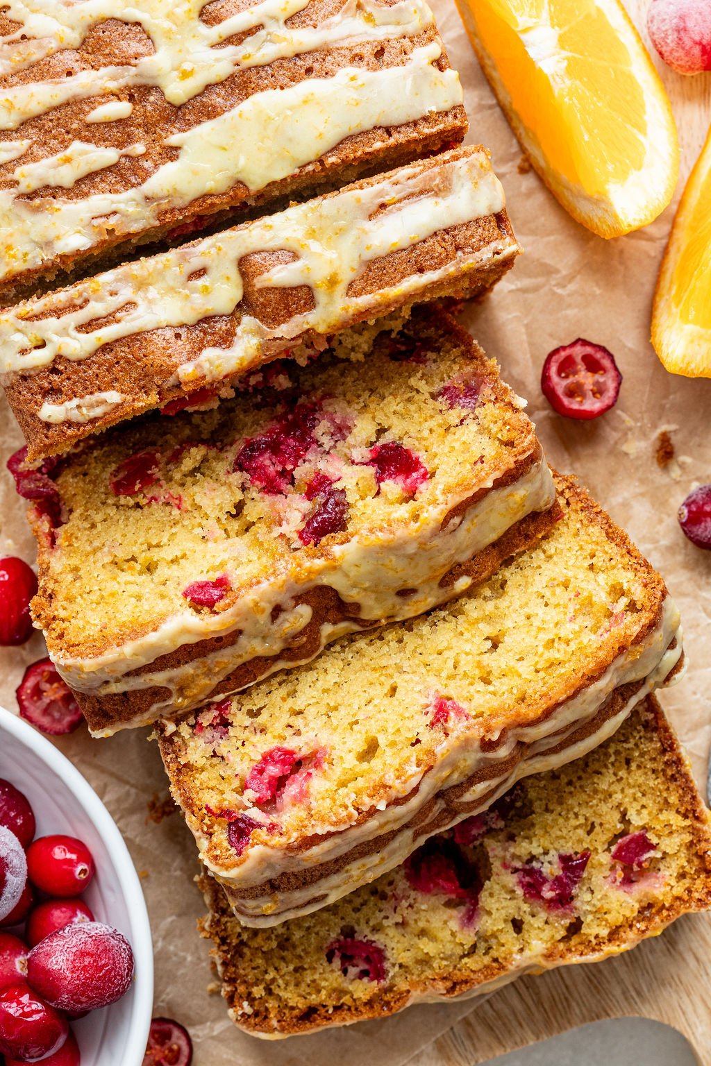 Sliced cranberry orange bread with icing drizzle, surrounded by fresh cranberries and orange wedges on parchment paper.