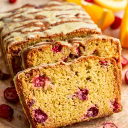A sliced loaf of cranberry orange bread with a light glaze, surrounded by fresh cranberries and orange wedges on a wooden surface.
