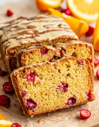 A sliced loaf of cranberry orange bread with a light glaze, surrounded by fresh cranberries and orange wedges on a wooden surface.