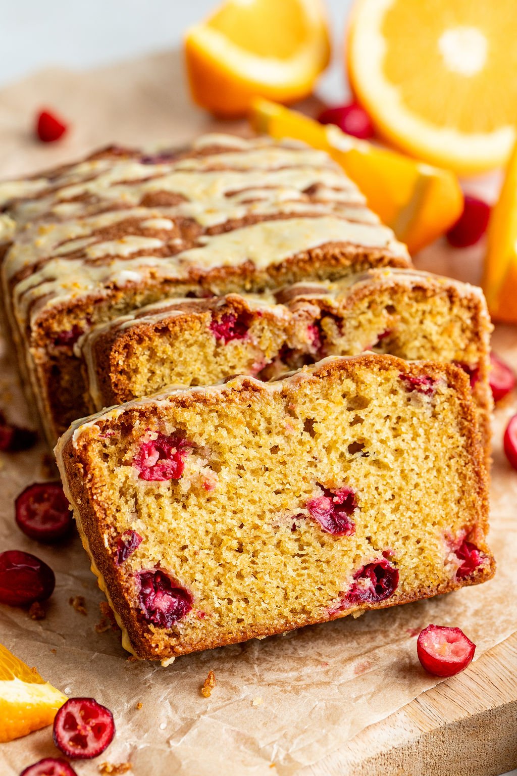 A sliced loaf of cranberry orange bread with a light glaze, surrounded by fresh cranberries and orange wedges on a wooden surface.