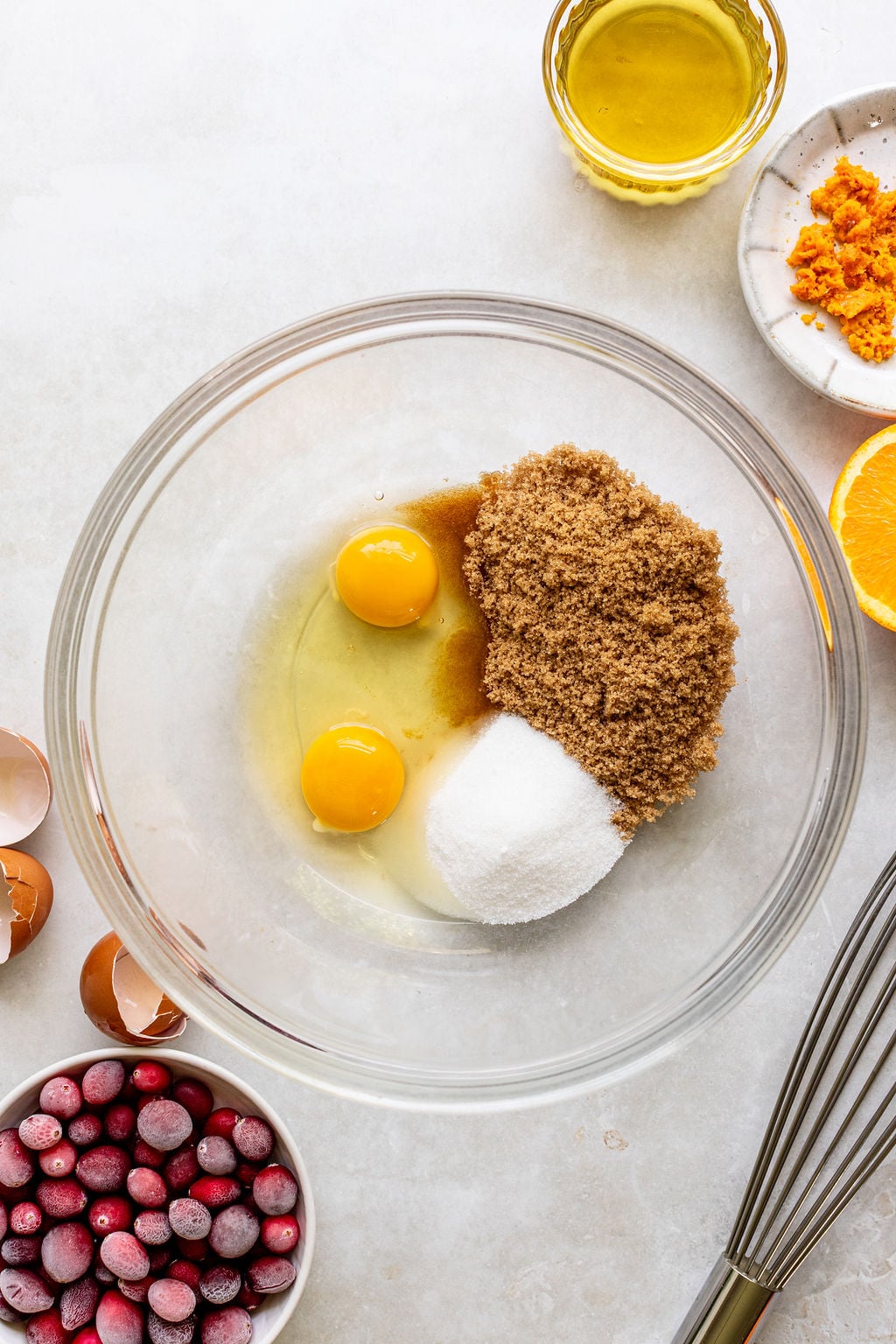 A glass bowl with two eggs, brown and white sugar, and vanilla extract. Surrounding are cranberries, orange zest, oil, eggshells, and a whisk on a white surface.