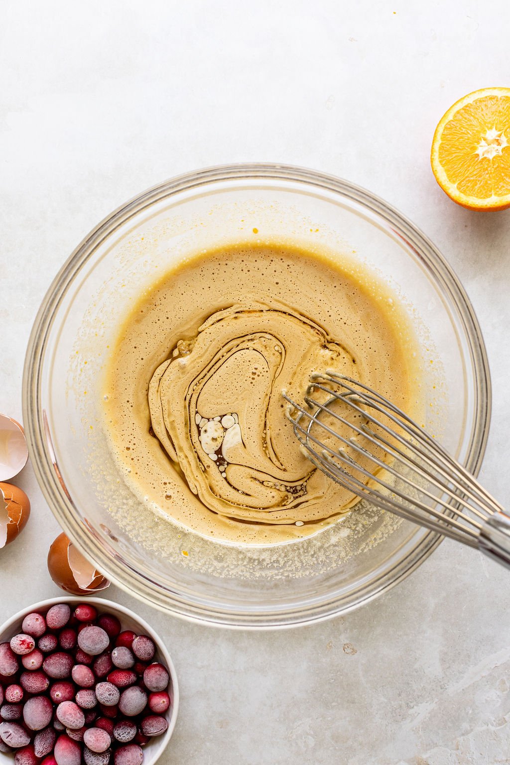 A glass bowl with a whisk mixing batter, surrounded by cracked eggshells, a halved orange, and a bowl of frozen cranberries on a light surface.