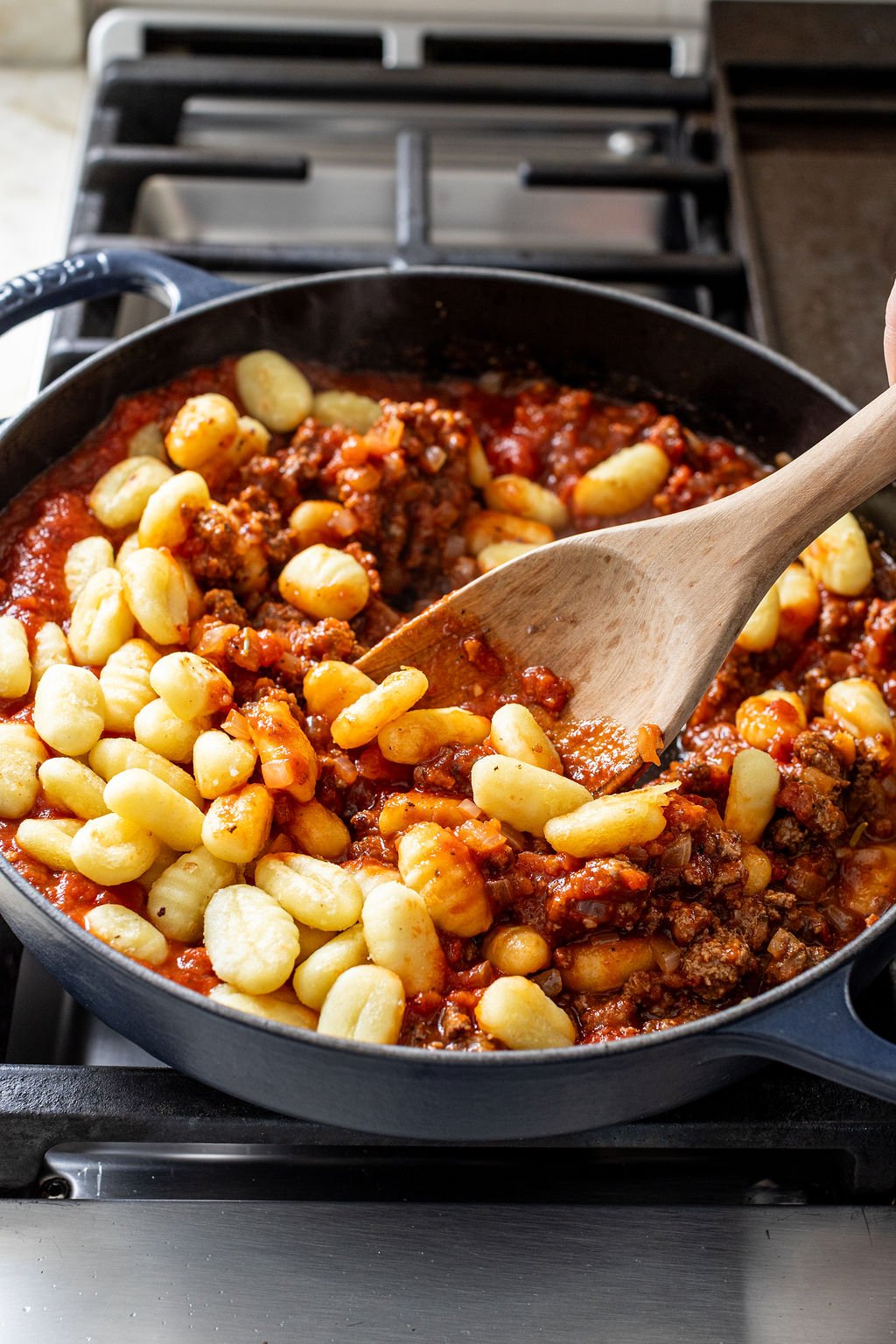 A wooden spoon stirs gnocchi with meat sauce in a black skillet on a stovetop.