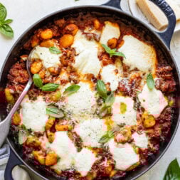 A cast iron skillet filled with baked gnocchi in tomato sauce, topped with melted mozzarella and fresh basil leaves, sits on a light countertop with a serving spoon.