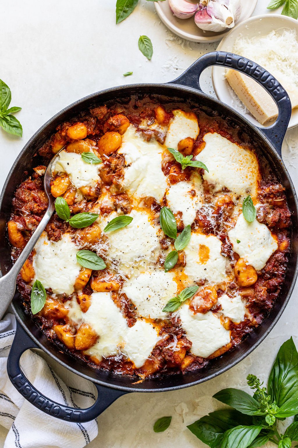 A cast iron skillet filled with baked gnocchi in tomato sauce, topped with melted mozzarella and fresh basil leaves, sits on a light countertop with a serving spoon.