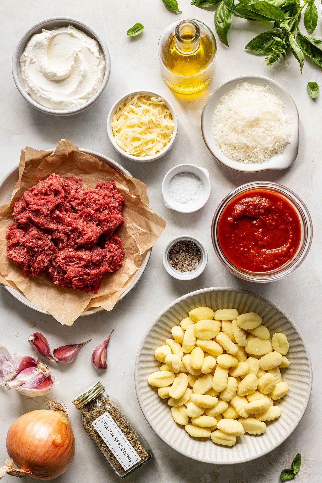 Ground beef, gnocchi, cheeses, tomato sauce, olive oil, herbs, garlic, onion, and seasonings on a table.
