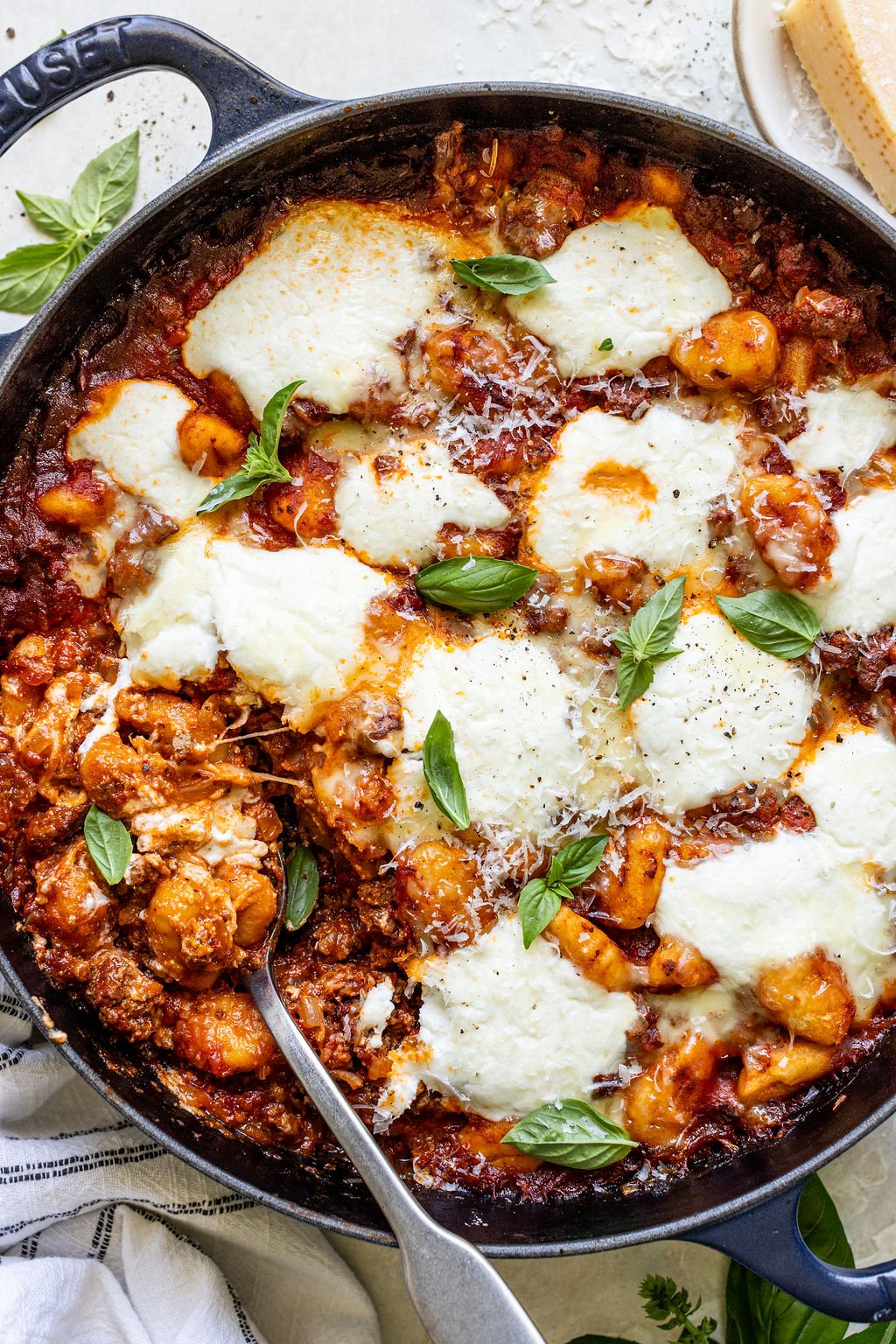 A skillet filled with baked gnocchi in tomato sauce, topped with melted cheese, fresh basil leaves, and a serving spoon.