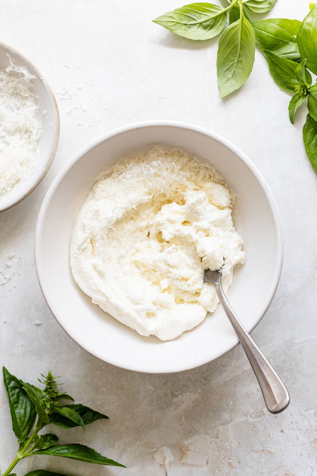 A white bowl with ricotta cheese and grated parmesan, partially mixed with a spoon, surrounded by fresh basil leaves on a light surface.