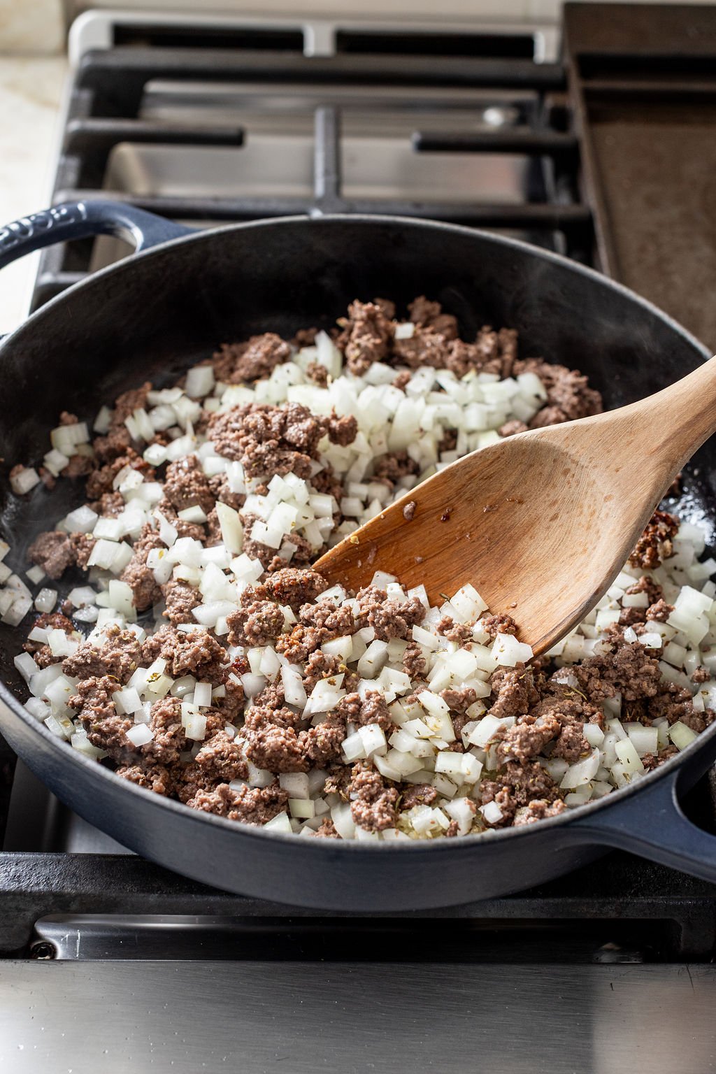 Ground beef and chopped onions being sautéed in a black skillet on a stovetop, stirred with a wooden spoon.