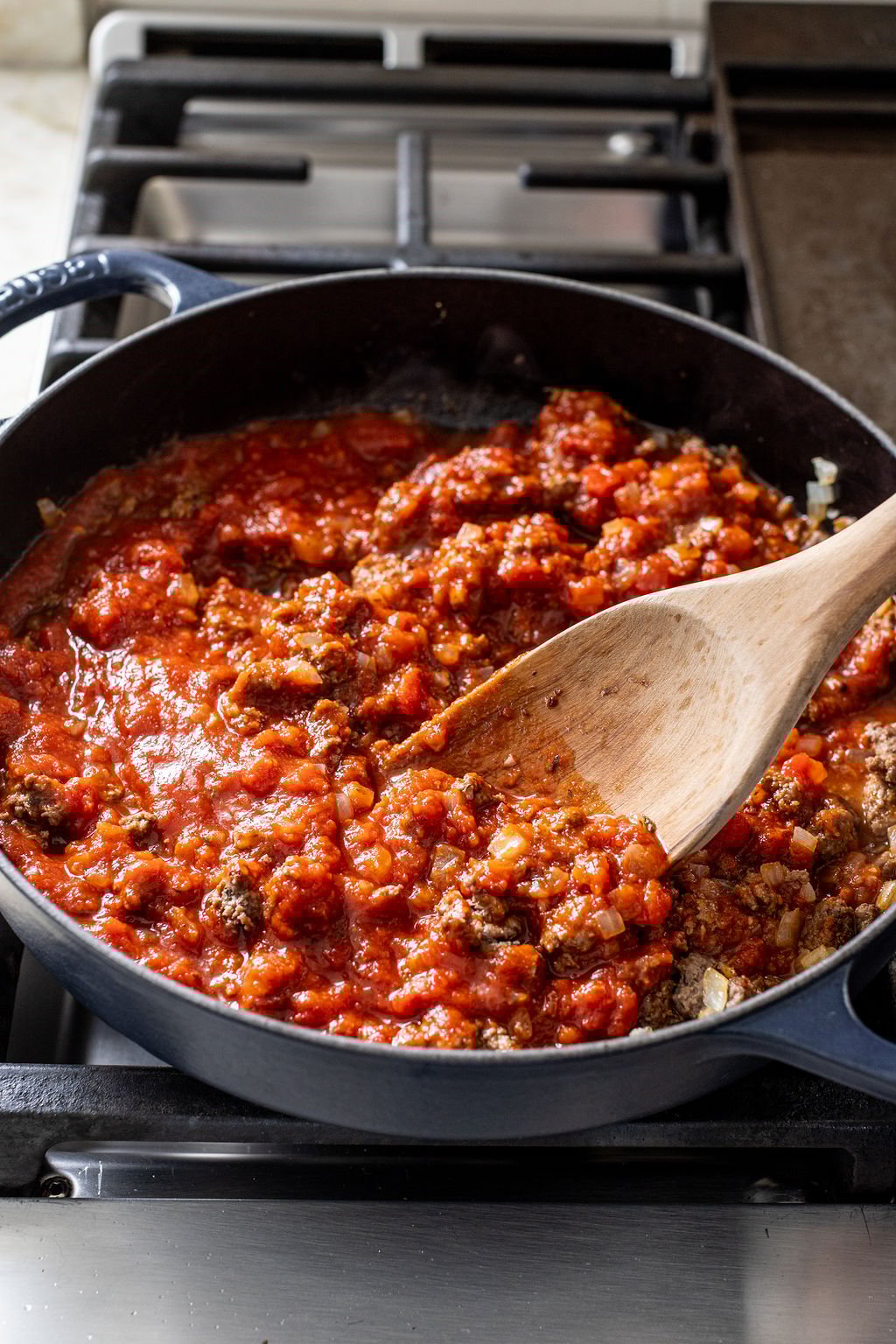 A skillet with tomato meat sauce being stirred with a wooden spoon on a stovetop.