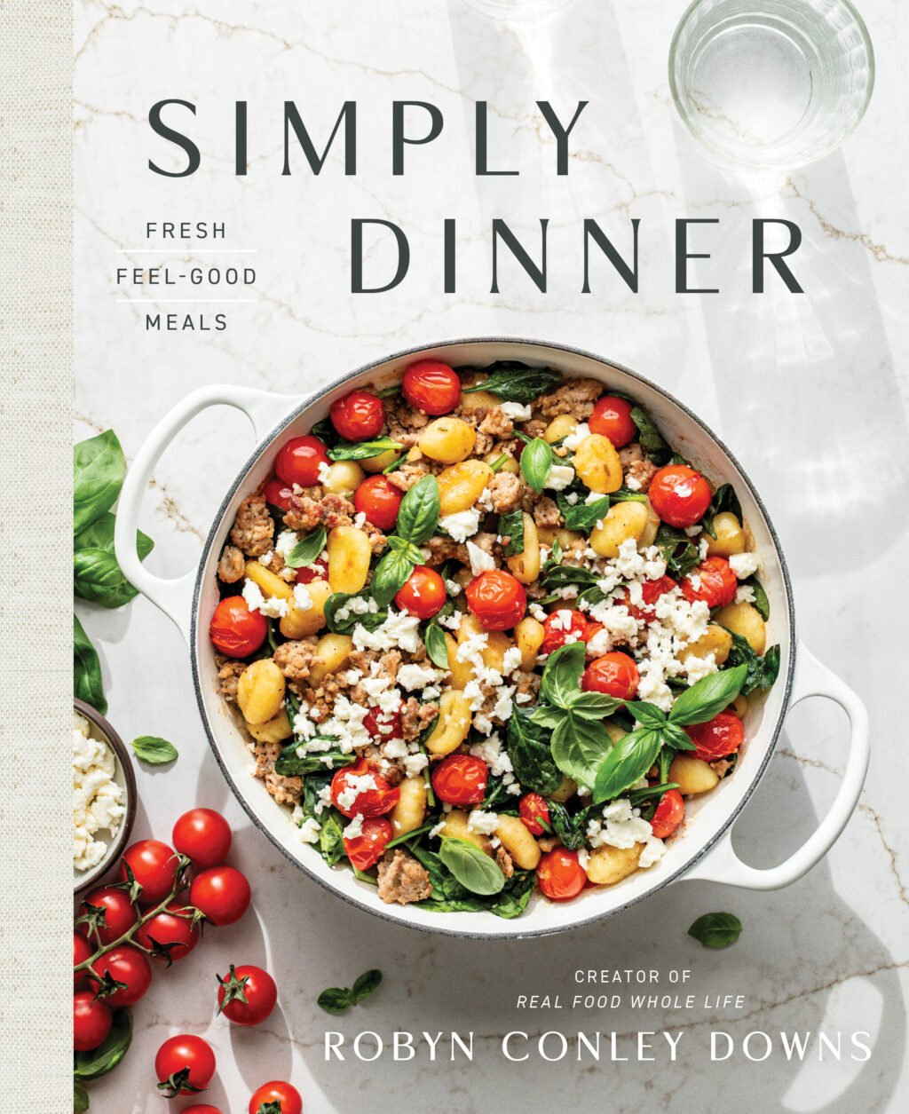 A cookbook titled Simply Dinner by Robyn Conley Downs, featuring a white pot of pasta with cherry tomatoes, greens, feta cheese, and herbs on a marble surface.