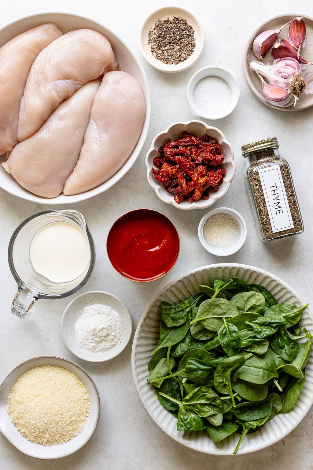 An overhead view of raw chicken breasts, baby spinach, sun-dried tomatoes, cream, tomato sauce, grated cheese, flour, garlic, dried thyme, salt, pepper, and seasonings on a white surface.