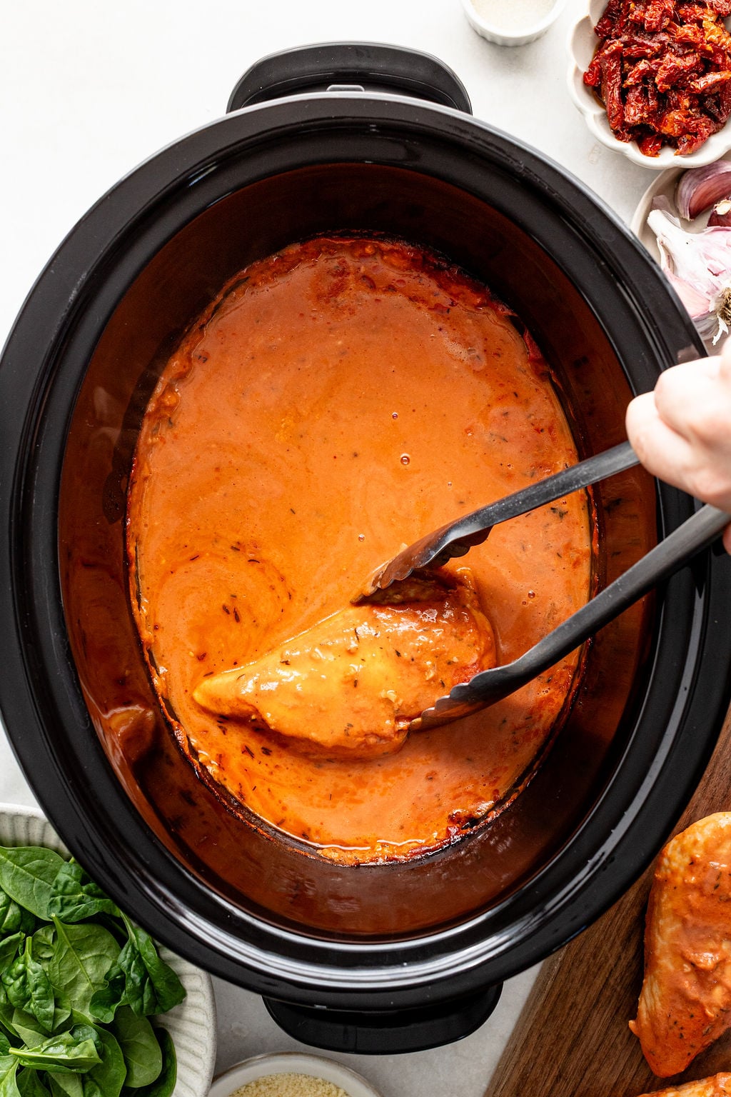 A hand uses tongs to lift a piece of chicken from a slow cooker filled with creamy orange sauce, surrounded by bowls of spinach, parmesan, sun-dried tomatoes, and shallots.