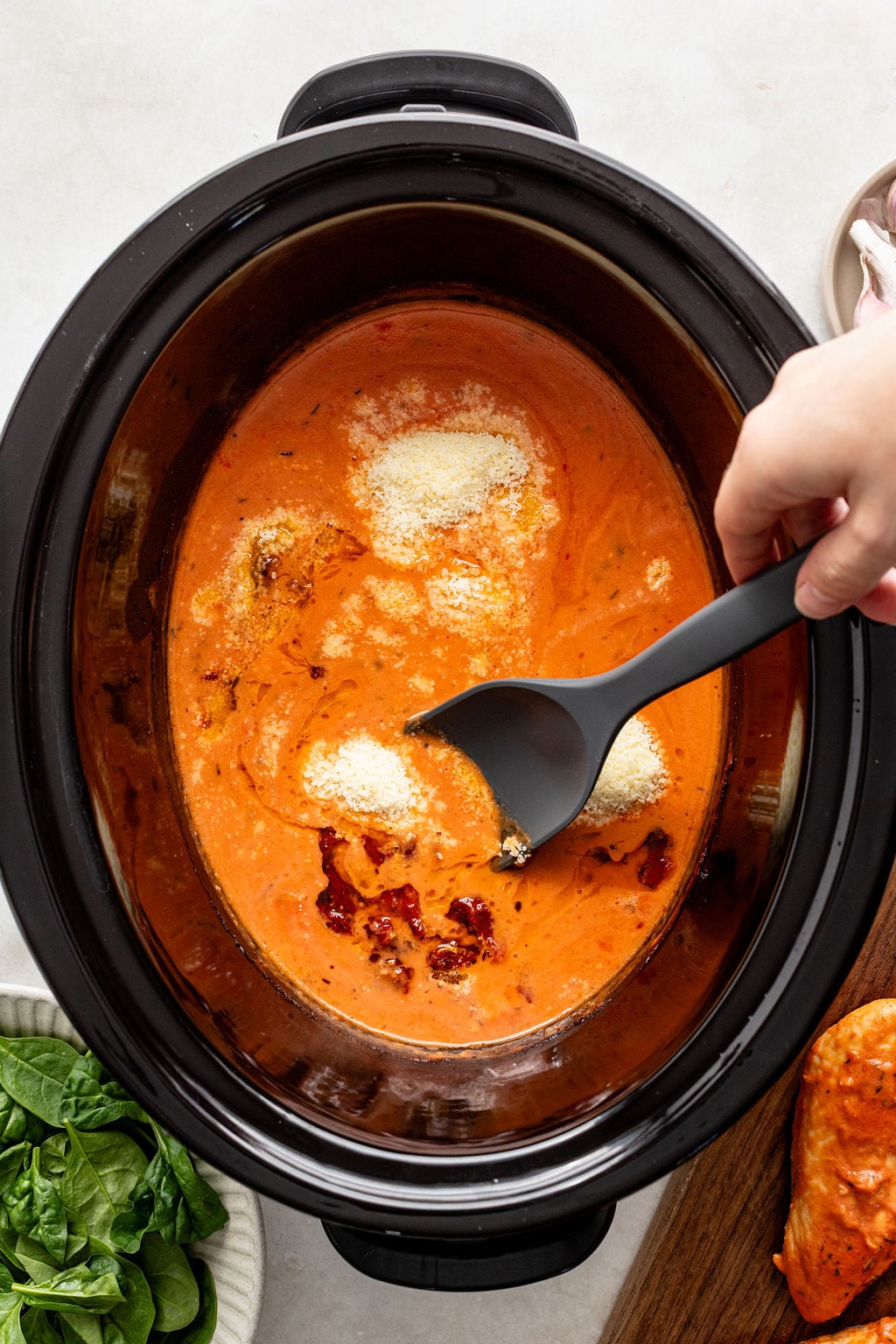A hand stirs a creamy tomato sauce with grated cheese in a slow cooker.