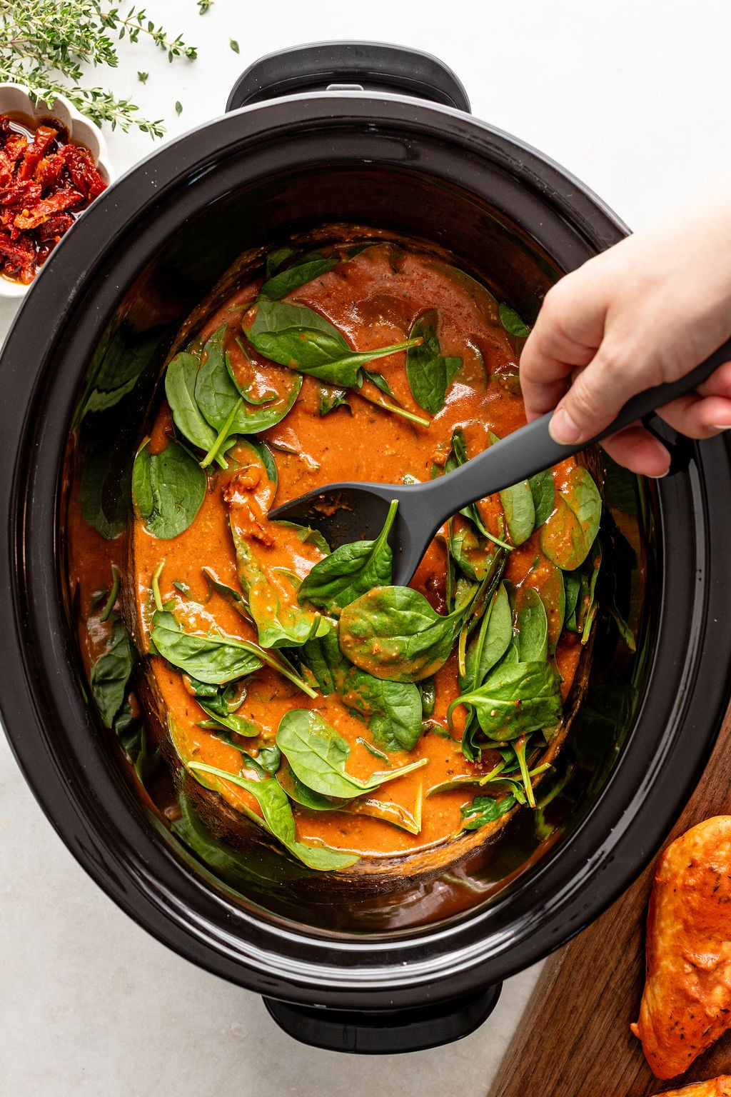 A hand stirs fresh spinach into a creamy red sauce with chicken in a slow cooker.