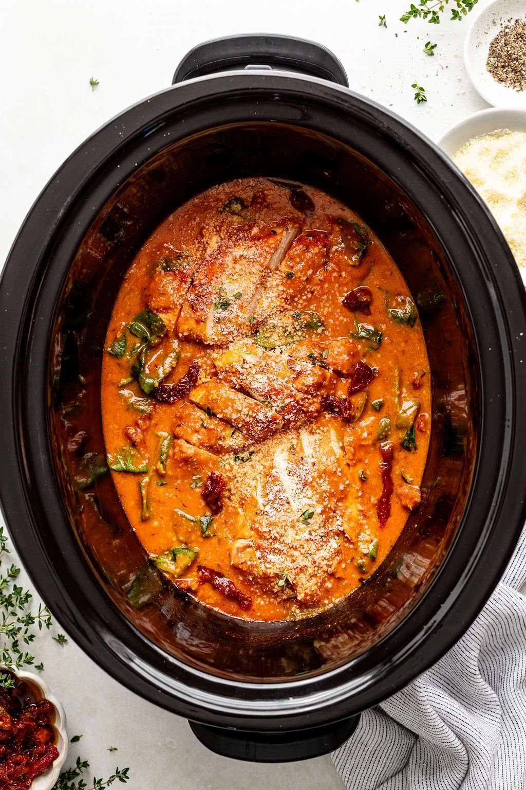 Slow cooker filled with chicken breasts in a creamy tomato sauce with spinach and sprinkled cheese, surrounded by small bowls of spices and herbs.