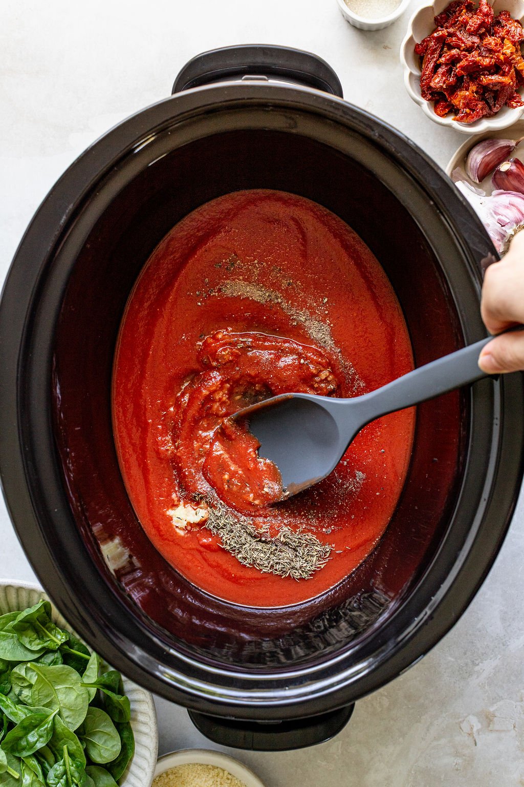 A hand stirs tomato sauce with herbs in a slow cooker.