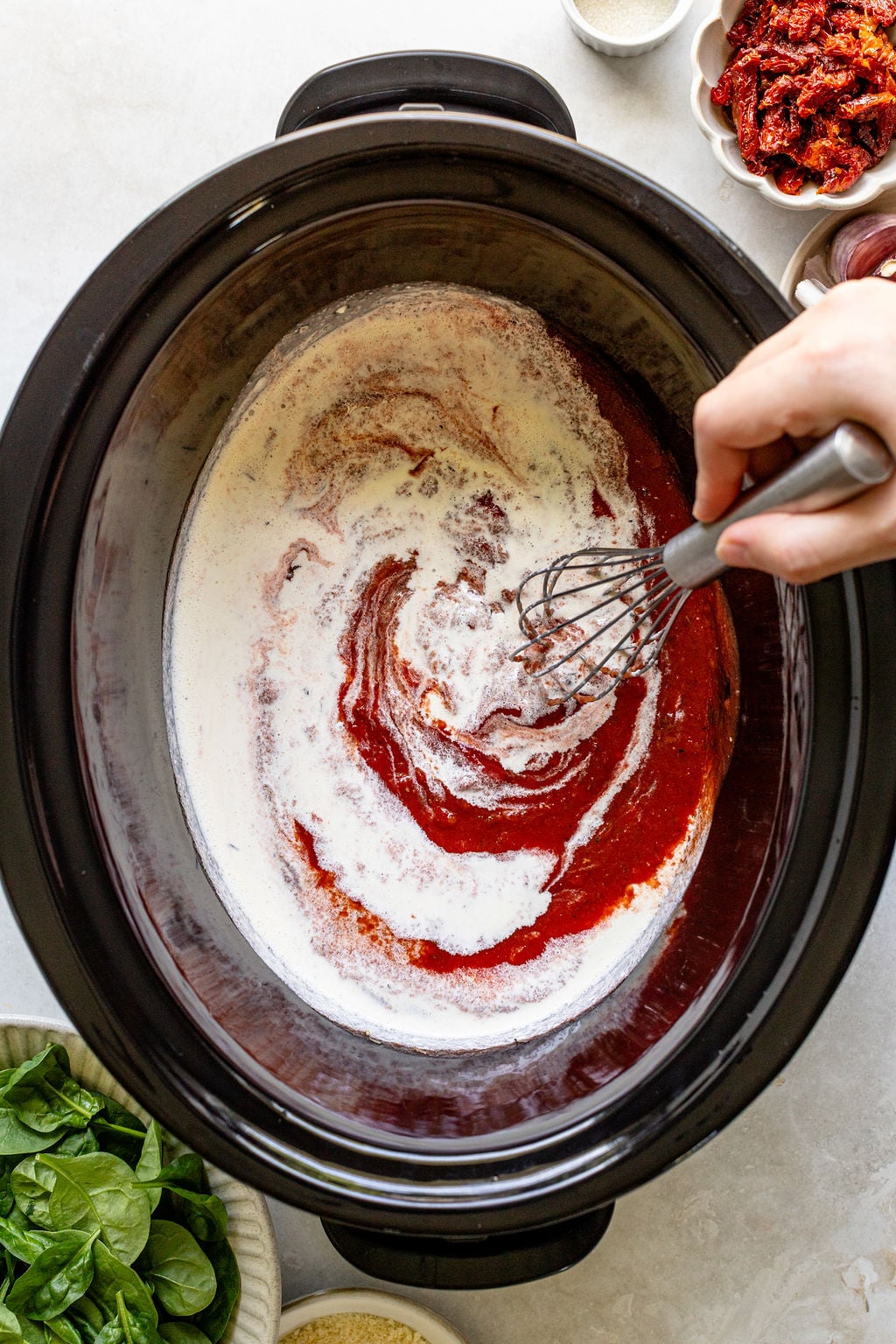A hand uses a whisk to mix cream and tomato sauce in a slow cooker.