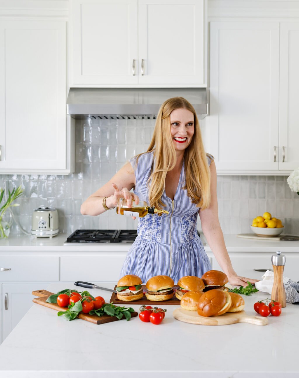 Robyn Conley Down in a blue dress prepares burgers in a bright kitchen, surrounded by fresh tomatoes, basil, and buns on a white countertop.