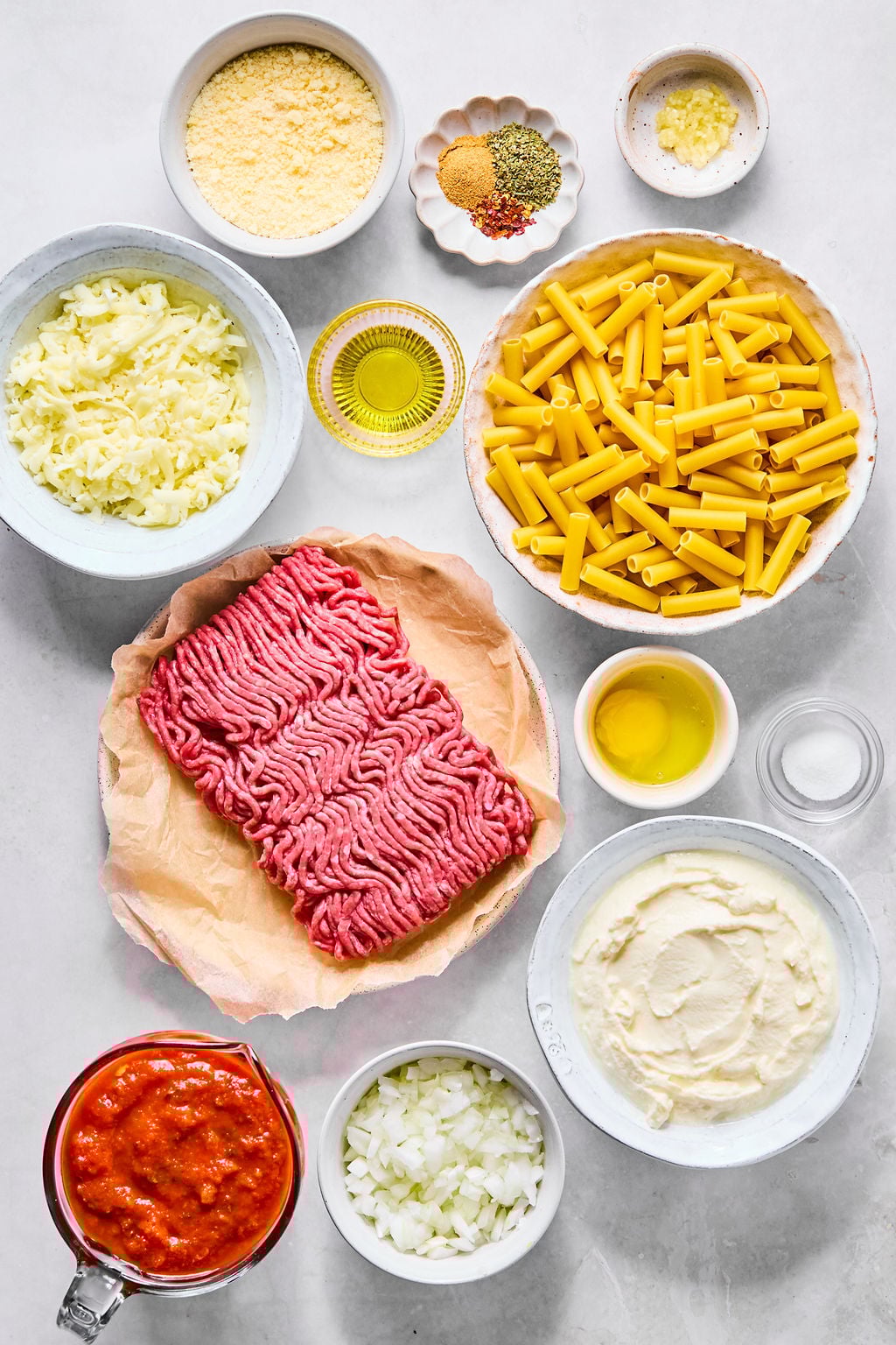 Ground meat, pasta, tomato sauce, cheese, chopped onions, spices, oil, and garlic on a marble counter.