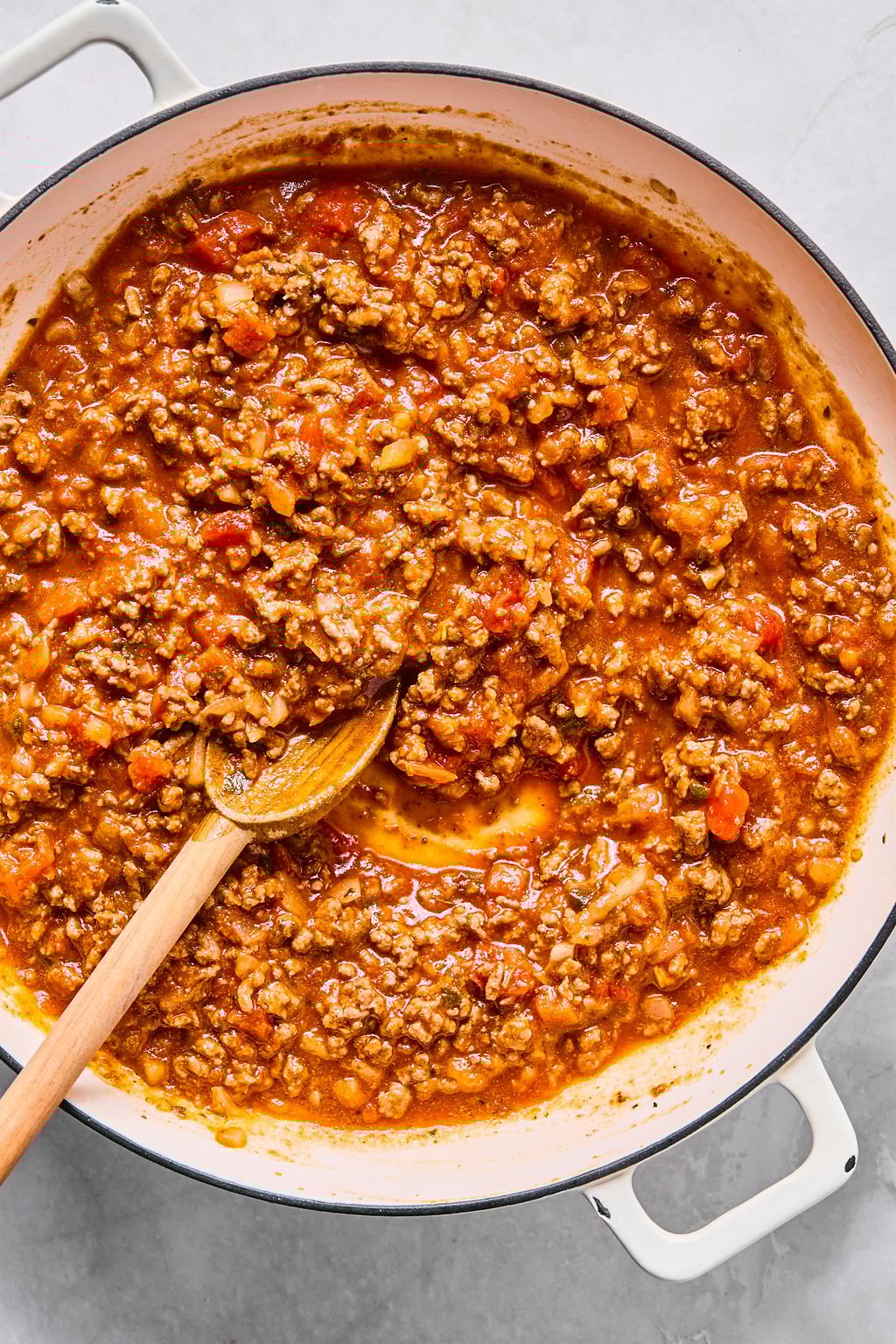 A white pot filled with cooked ground meat in tomato sauce is being stirred with a wooden spoon.