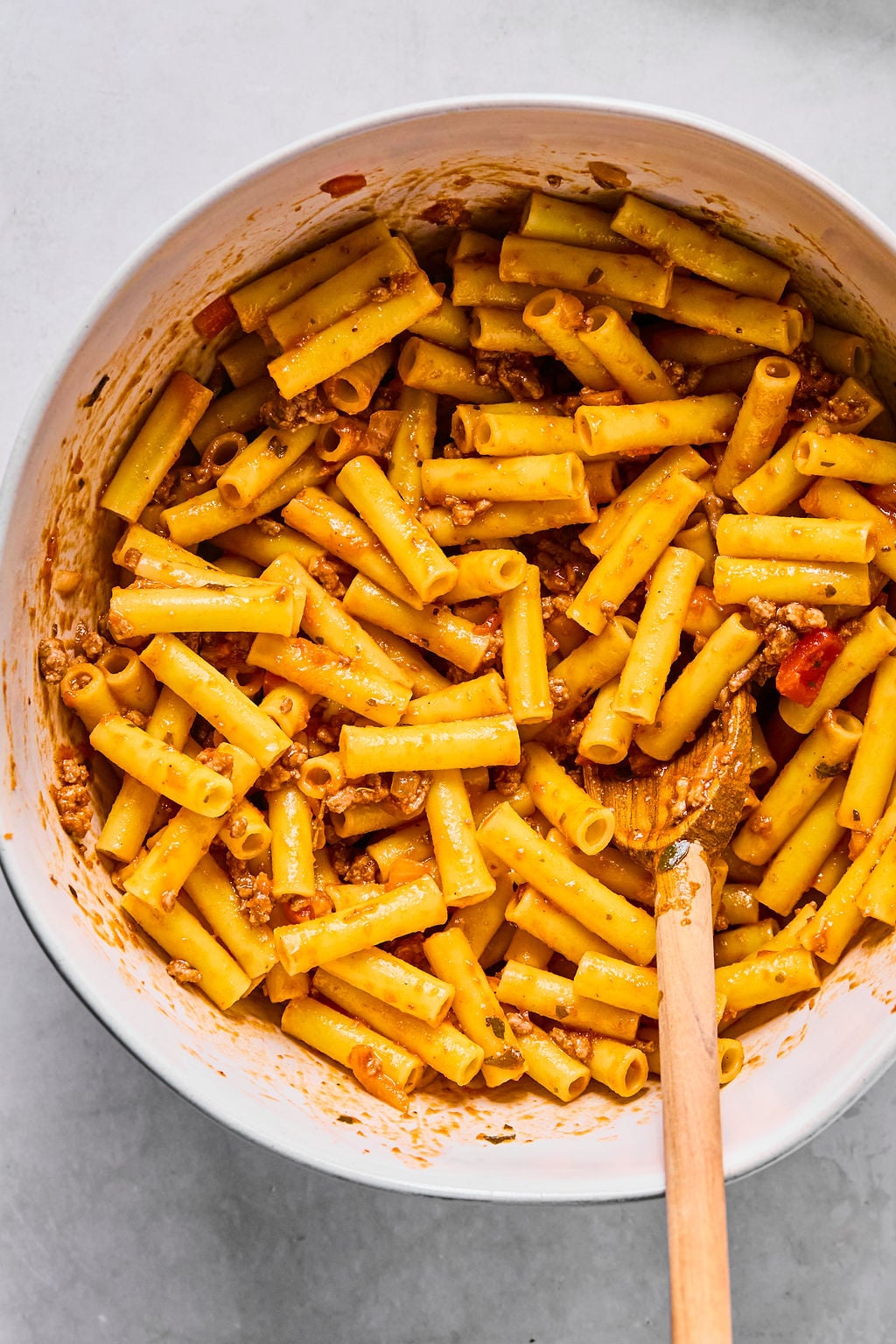 A bowl of cooked ziti pasta mixed with tomato sauce and ground meat, stirred with a wooden spoon.