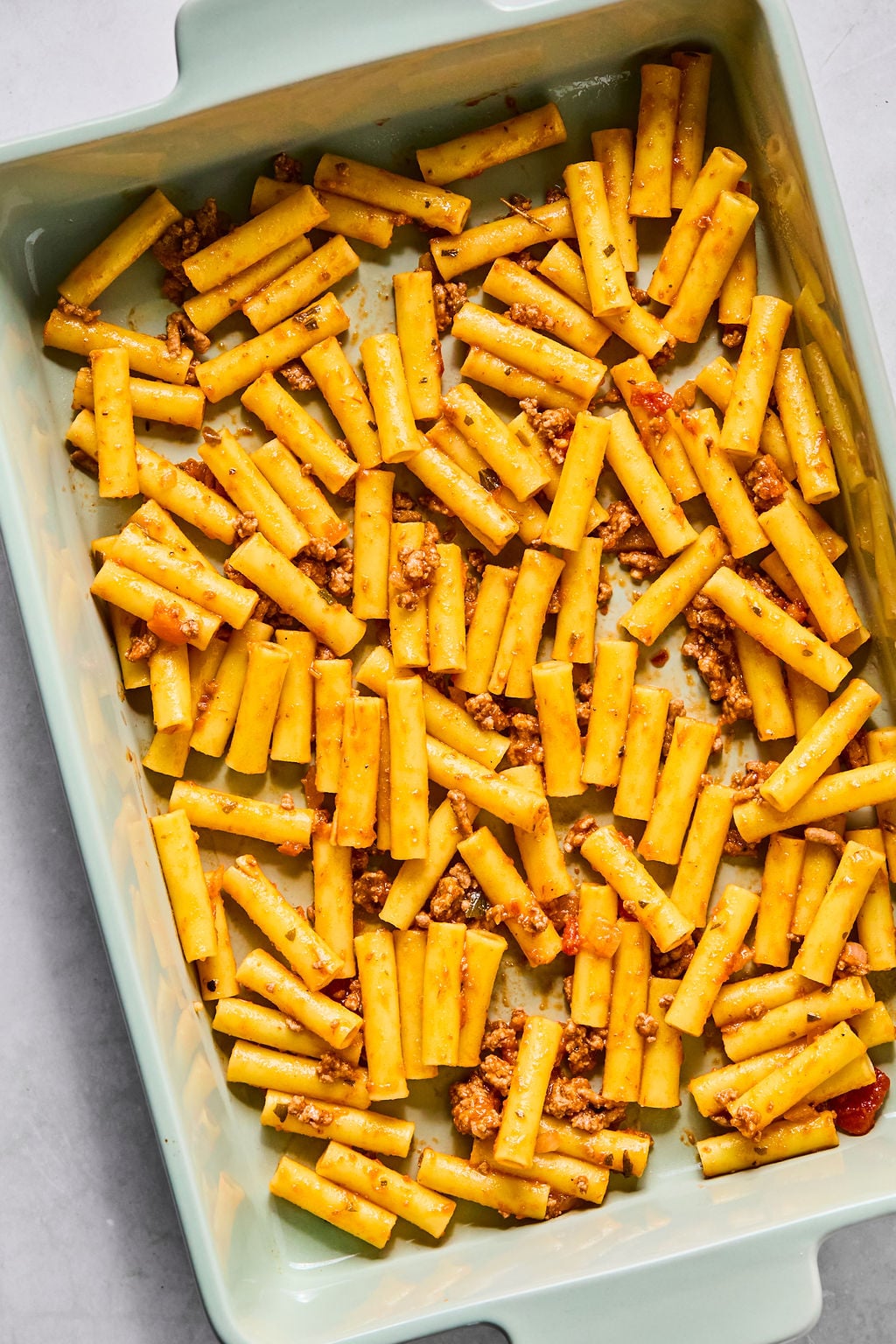 Cooked ziti pasta mixed with ground meat and tomato sauce spread in a rectangular baking dish.