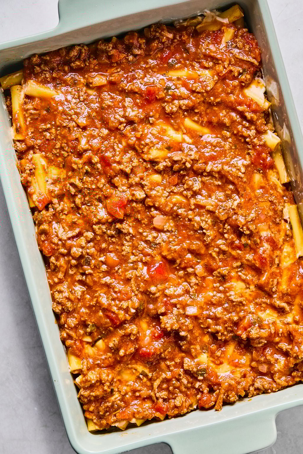 A baking dish filled with uncooked lasagna, topped with a layer of meat sauce containing ground beef and diced tomatoes.