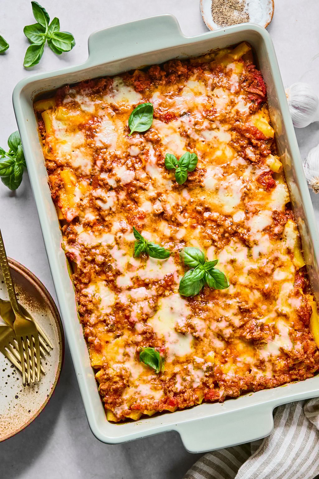 A baked casserole dish filled with cheesy ziti topped with tomato sauce, ground meat, melted cheese, and fresh basil leaves, shown alongside plates, forks, and seasonings.
