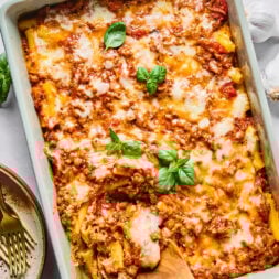A baked pasta dish with melted cheese and tomato meat sauce, garnished with fresh basil leaves, in a rectangular pan with a wooden spoon inside.