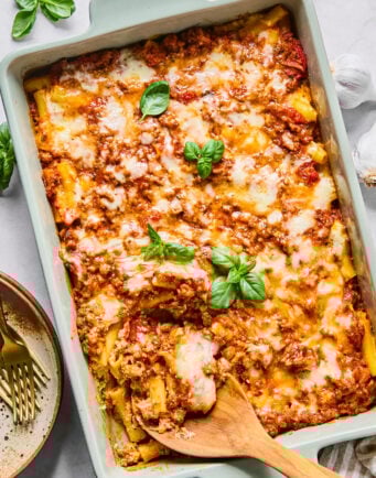 A baked pasta dish with melted cheese and tomato meat sauce, garnished with fresh basil leaves, in a rectangular pan with a wooden spoon inside.