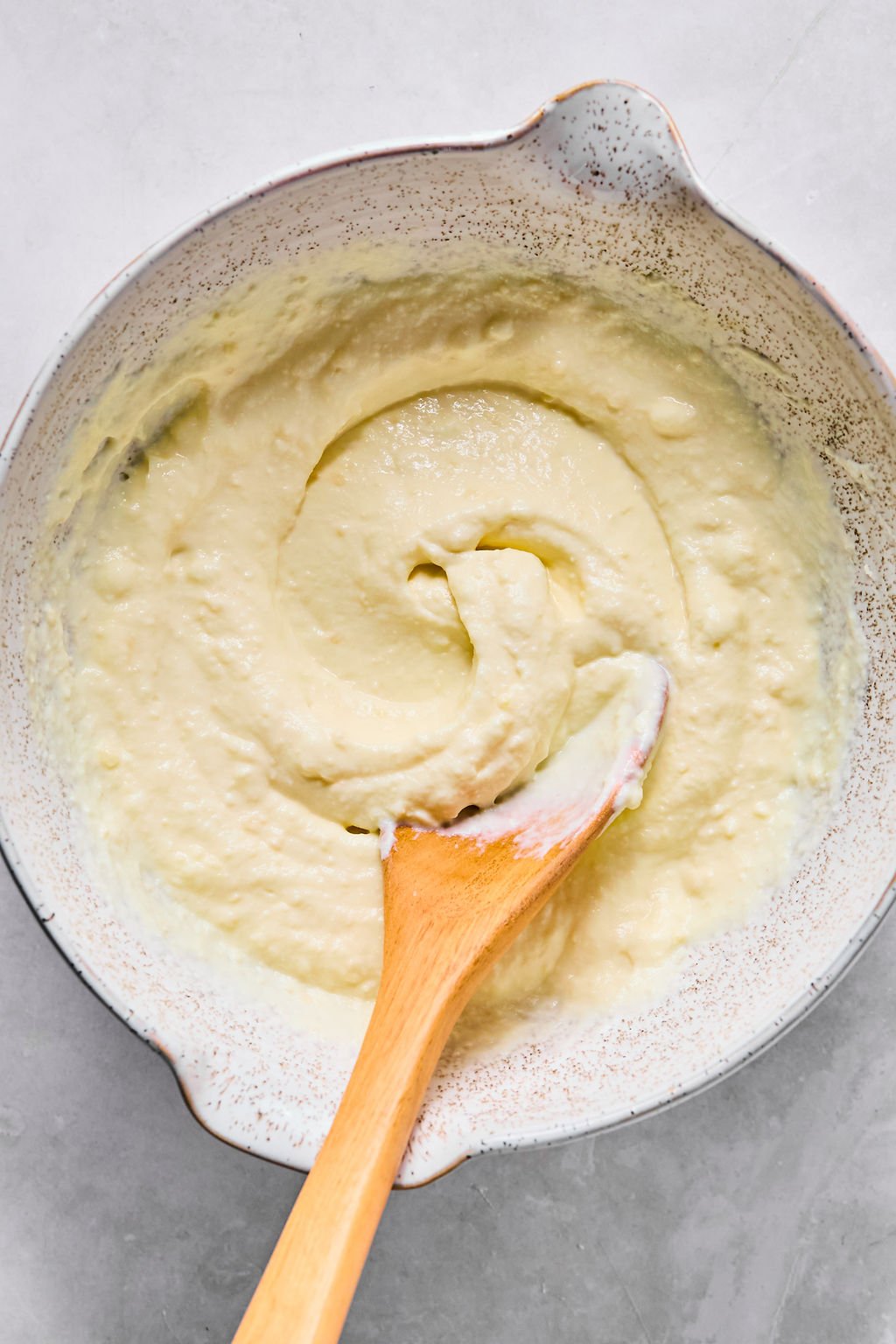 A ceramic bowl filled with thick, creamy ricotta mixture being stirred with a wooden spoon on a light countertop.