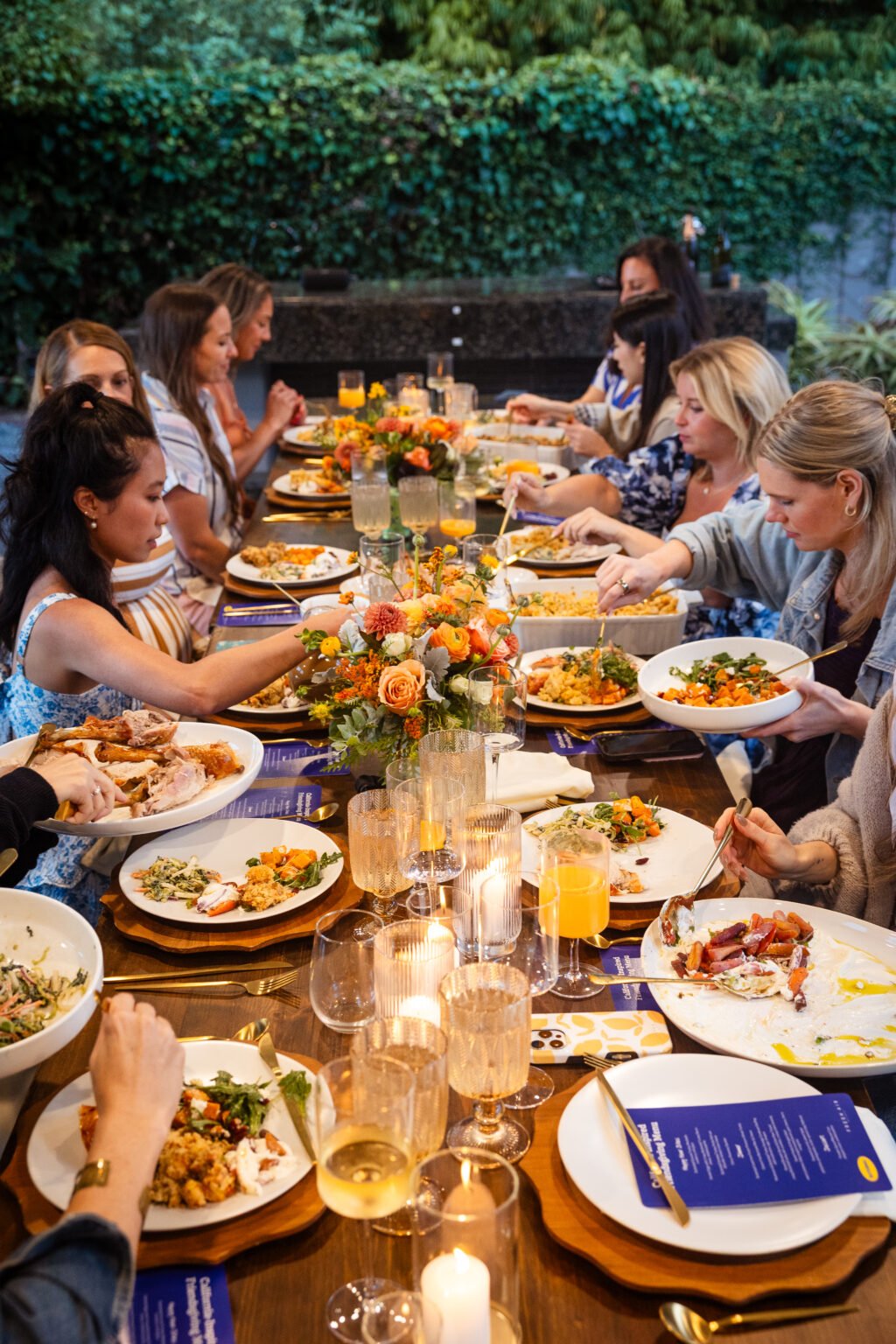 A wooden table is set for a formal meal with white plates, blue menus, gold cutlery, crystal glasses, and orange floral centerpieces.