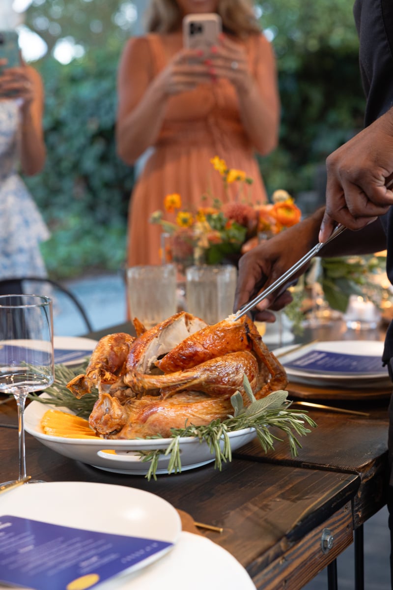 Person carving a roasted turkey at a dinner table set with plates, glasses, and floral centerpiece, while others stand in the background taking photos.