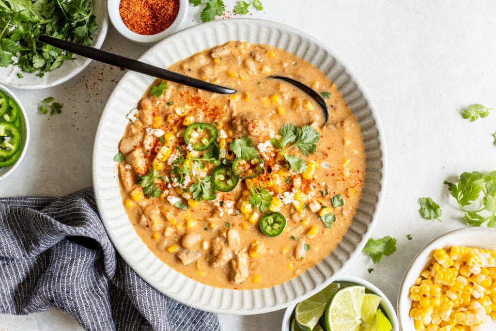 A bowl of creamy corn soup garnished with sliced jalapeños, cilantro, and crumbled cheese, surrounded by small dishes of lime wedges, corn, cilantro, and seasoning.