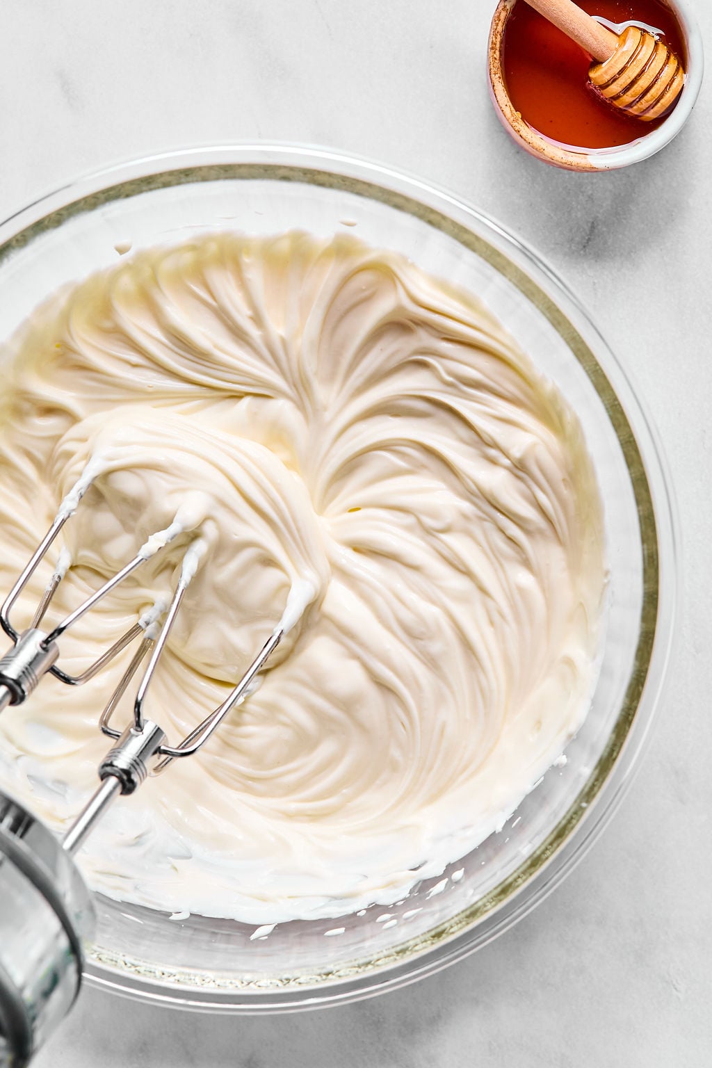 A bowl of whipped cream cheese being mixed with an electric mixer, with a small dish of honey and a honey dipper nearby on a white surface.