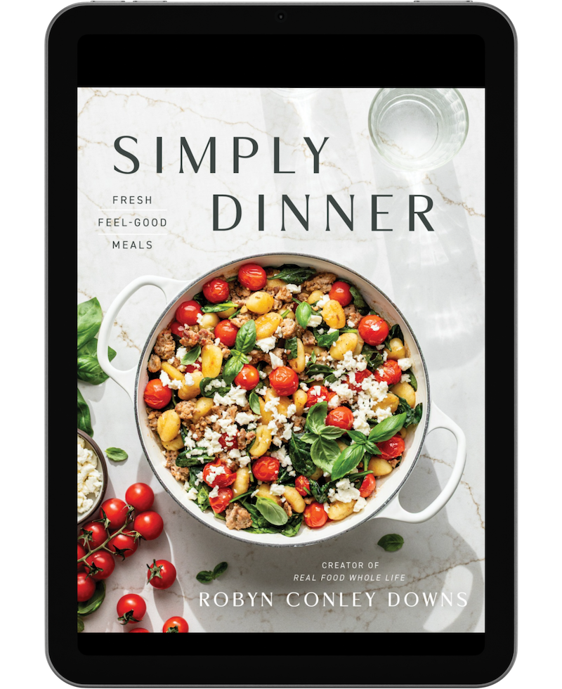 A tablet displays the cover of the cookbook Simply Dinner by Robyn Conley Downs, featuring a vegetable and grain dish in a white pot with scattered tomatoes and greens.