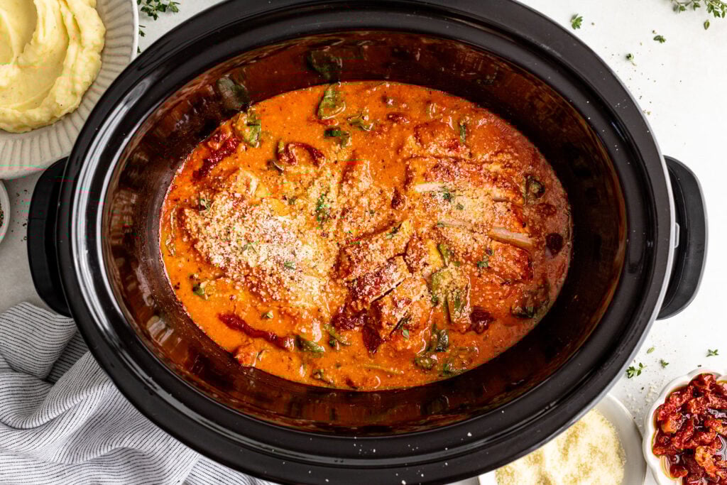 A slow cooker filled with creamy tomato-based chicken topped with grated cheese and herbs.
