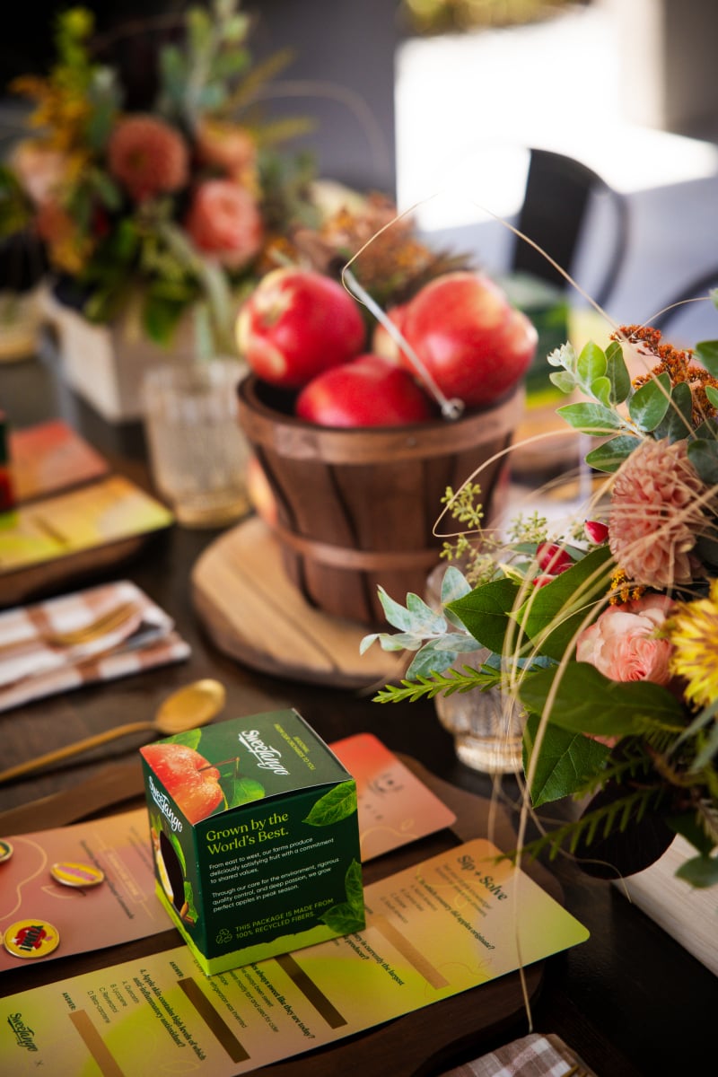 A table setting with a box of green tea, a wooden basket of apples, fresh flowers, and colorful place cards arranged neatly.
