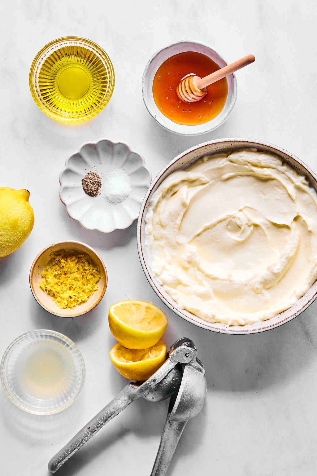 A bowl of creamy ricotta surrounded by bowls of olive oil, honey, zest, salt, pepper, a halved lemon, lemon juice, and a citrus press on a white surface.