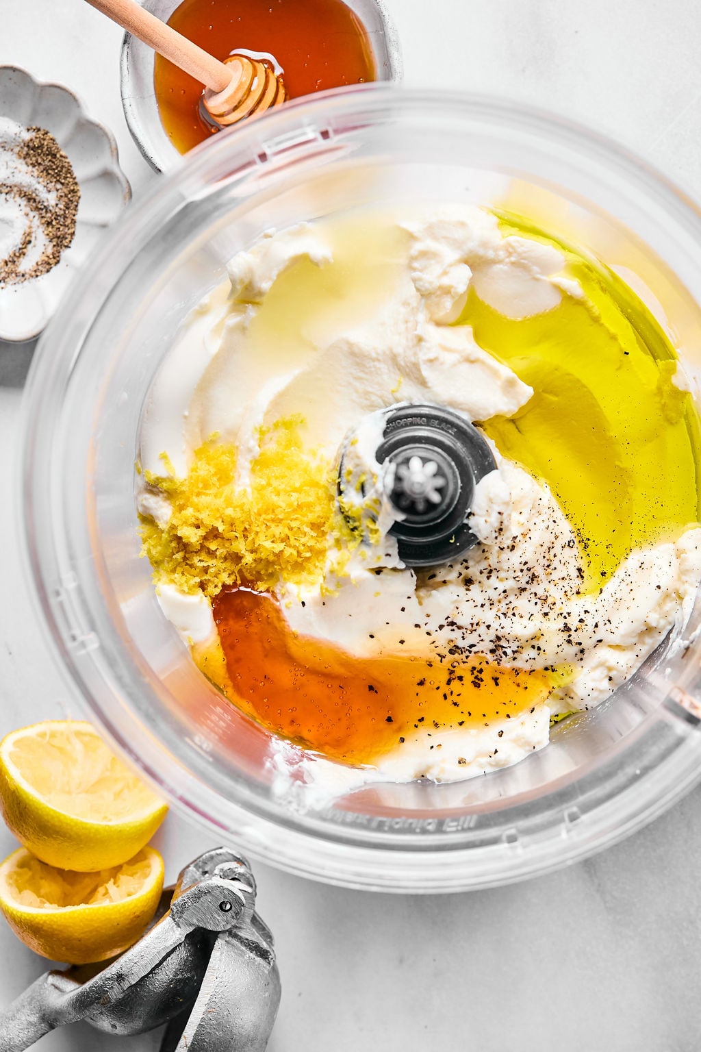 Top view of a food processor bowl containing ricotta, olive oil, honey, lemon zest, lemon juice, salt, and pepper, with a juicer, halved lemons, honey, and pepper nearby.