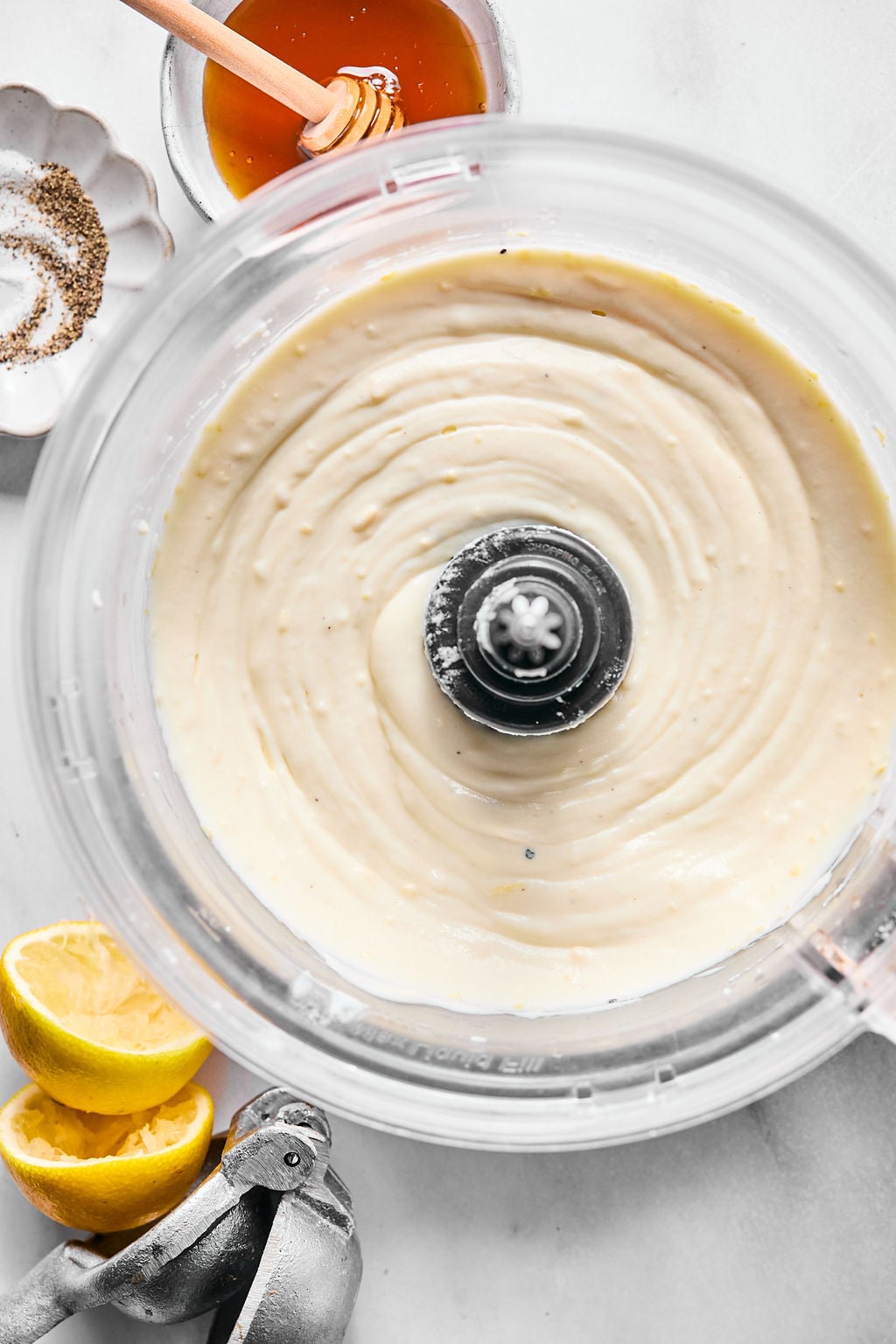 Overhead view of a food processor containing a creamy mixture, surrounded by a bowl of honey, a dish of salt, squeezed lemon halves, and a lemon squeezer on a white surface.